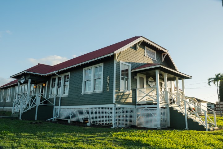 Old grey building with red roof and white trim, surrounded by grass, under a clear blue sky.