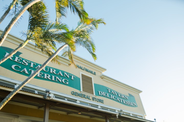 Building with signs: Restaurant Catering, Tavern Delicatessen, palm trees in foreground.