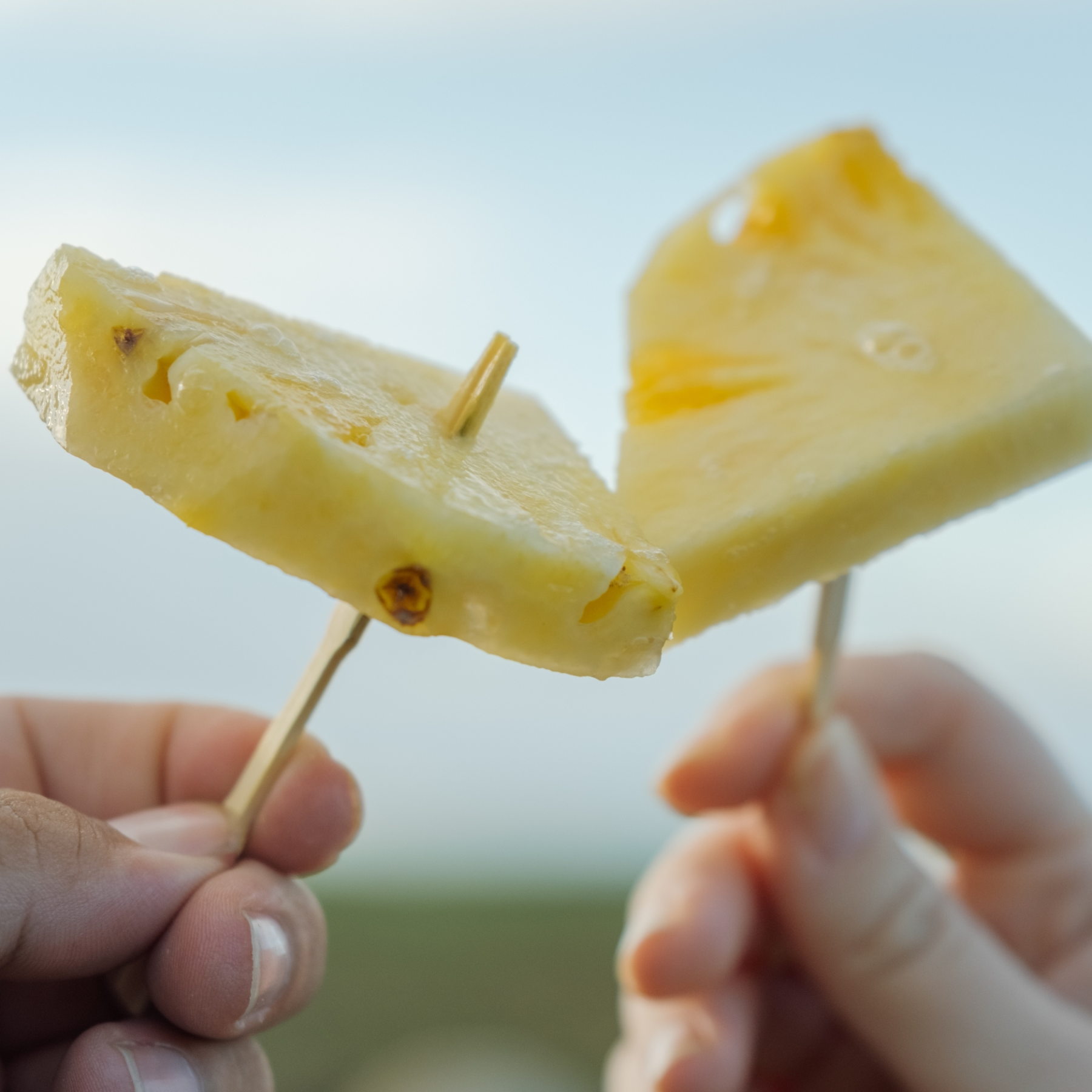 Two hands holding pineapple slices on sticks with a sky background.