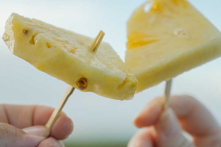 Two hands holding pineapple slices on sticks with a sky background.