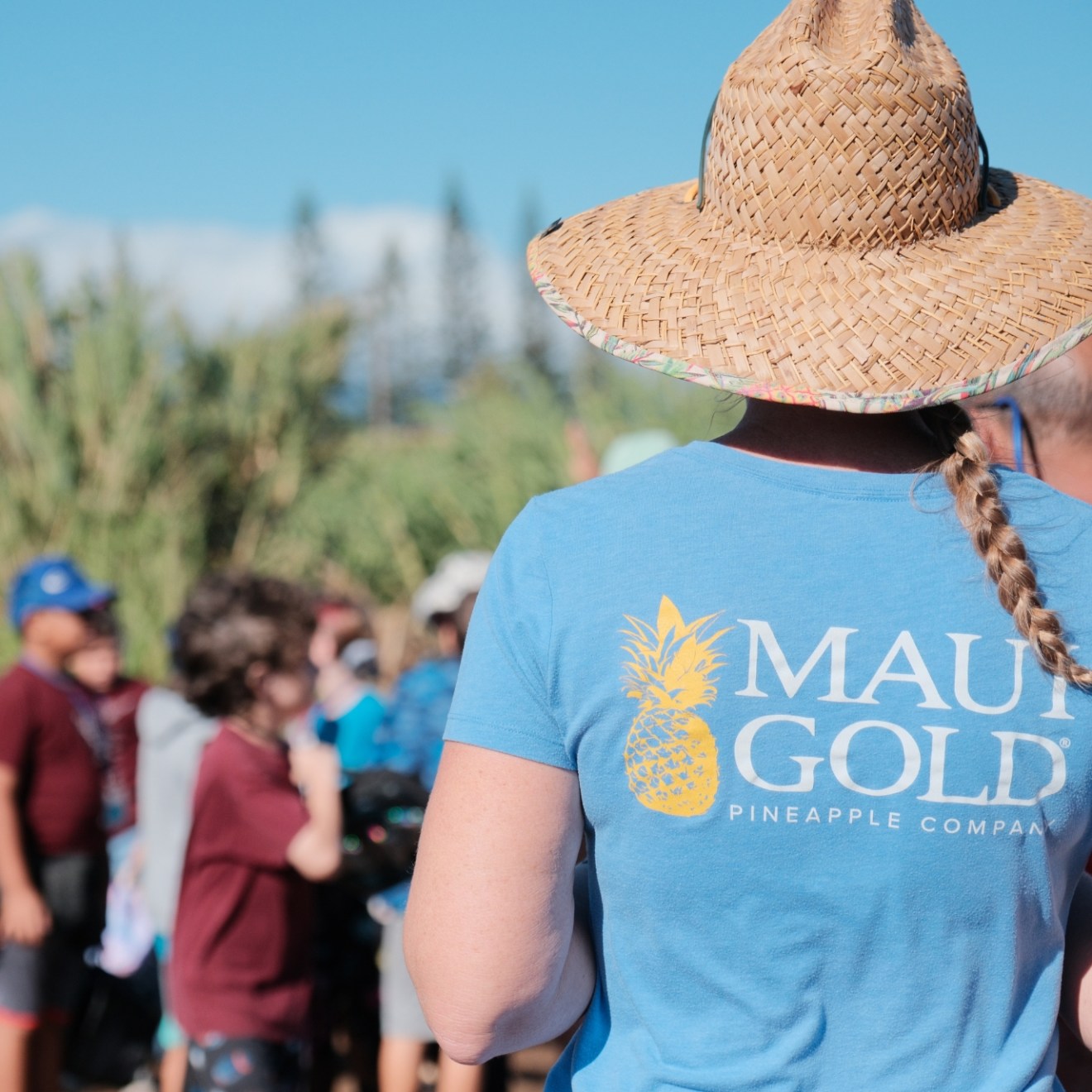 Person with straw hat and braid, wearing 'Maui Gold Pineapple Company' shirt, in a crowd outdoors.
